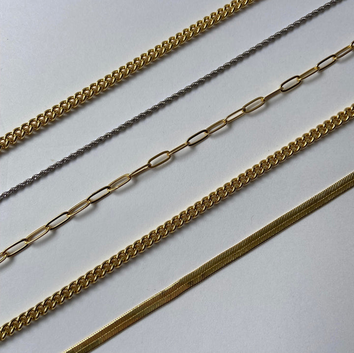 Multiple chains of different styles lined up diagonally for an up close shot. From left to right: gold curb chain, white gold rope chain, gold paperclip chain, gold curb chain and gold herringbone chain.